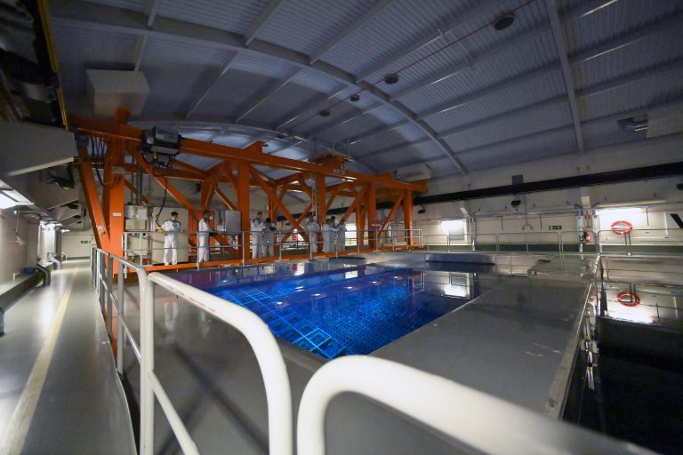 An image of blue water within a spent fuel pond inside a large building. Inside the pond, mesh racks of fuel rods are visible. A fuel-handling crane superstructure sits above the pool.