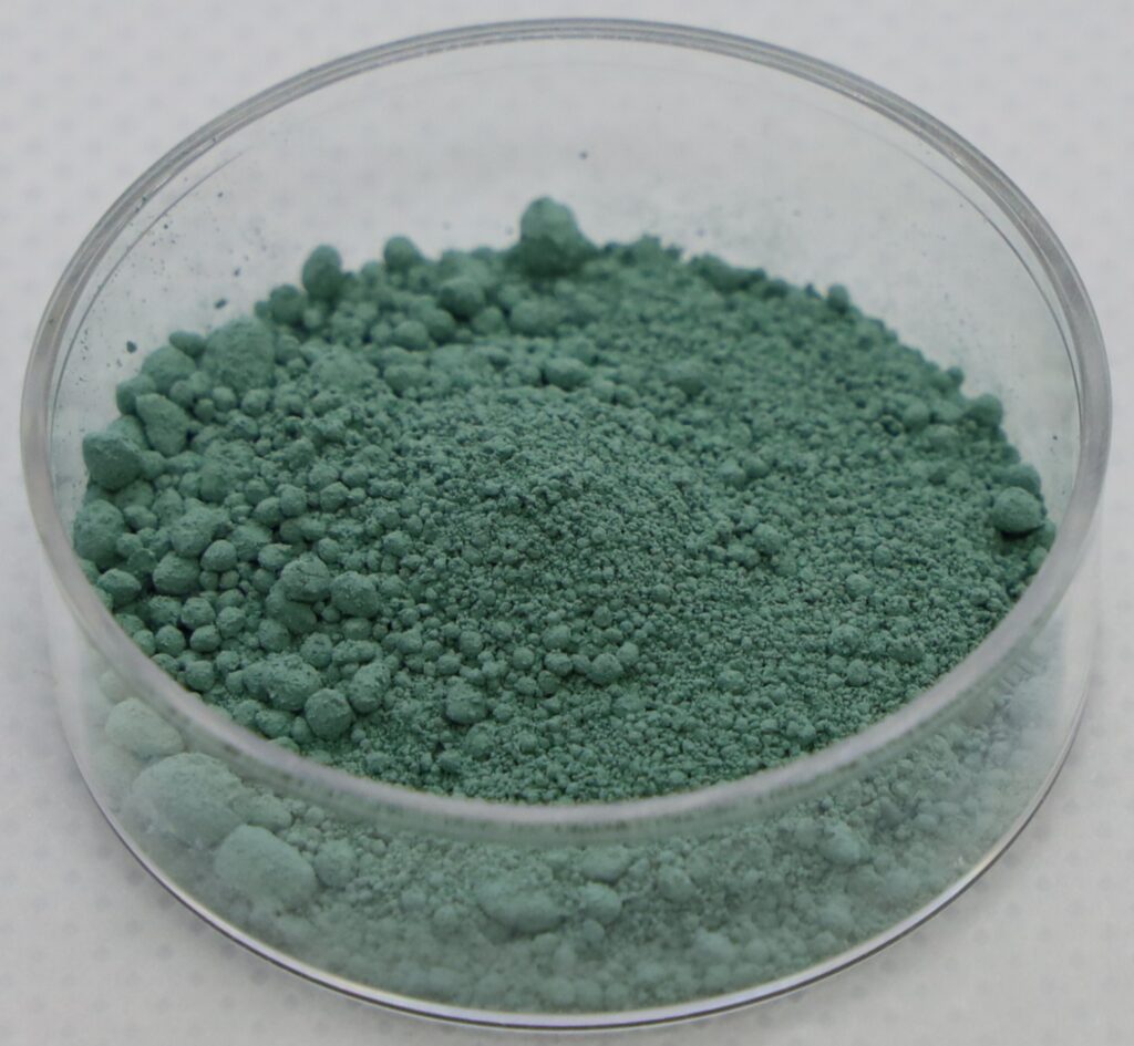 A glass dish containing green uranium tetrafluoride (UF₄) powder, a key intermediate compound in the nuclear fuel conversion process.